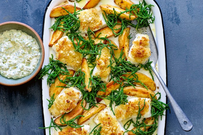 An oven tray filled with golden chunky chips and breadcrumbed hunks of fish on a blue background with a creamy dip