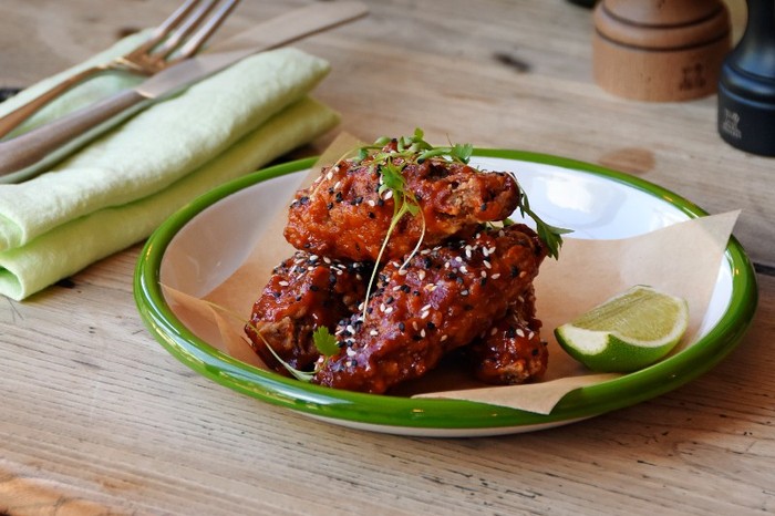 A white plate of Buttermilk-fried chicken wings with chilli and sesame next to cutery on a wooden table