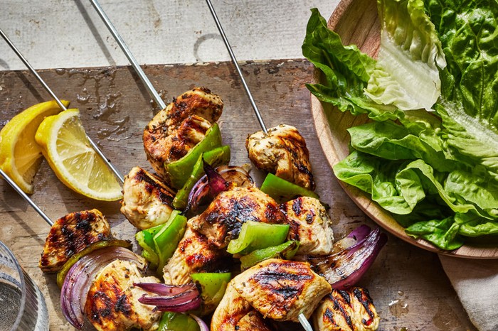 Chicken souvlaki skewers on a board, with a bowl of salad leaves on the side and two torn grilled pitta breads