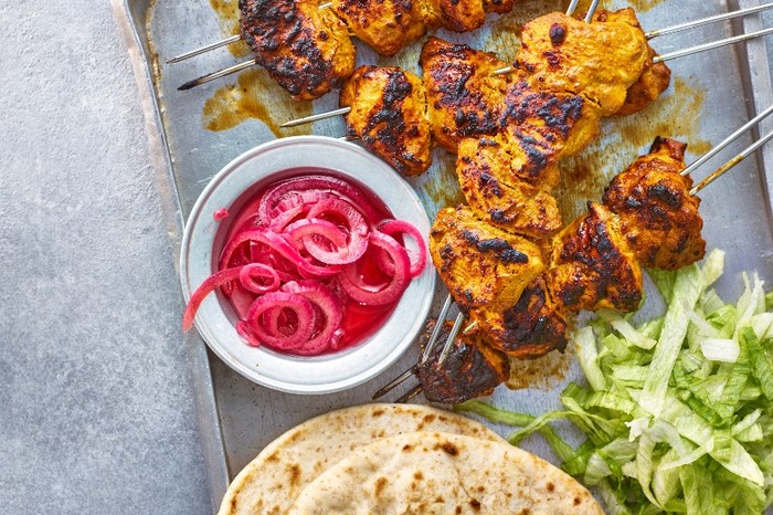 A tray of golden spiced chicken pieces on metal skewers with a bowl of pink pickled onions
