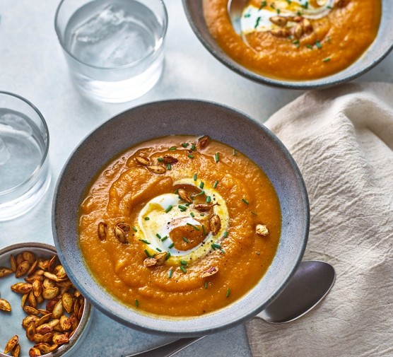 A dark grey bowl filled with orange soup with a swirl of yoghurt and toasted pumpkin seeds