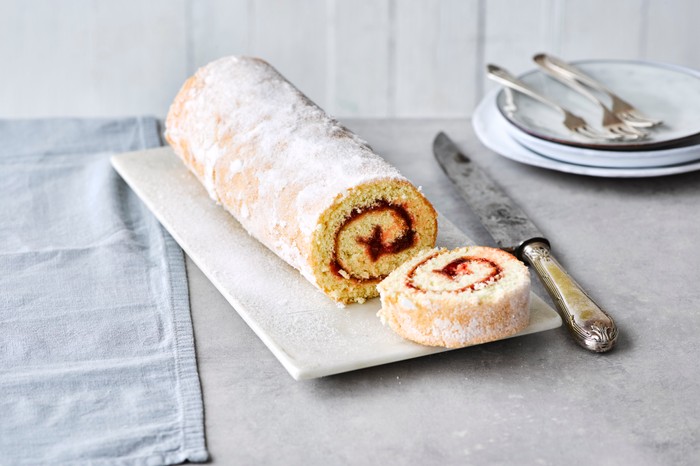 A swiss roll on a white board with empty side plates on the side and a dessert knife