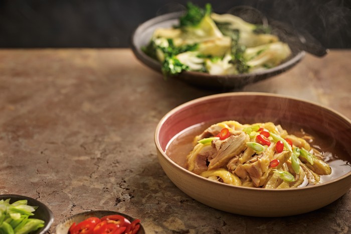 A terracotta bowl filled with pieces of steamed chicken in a chicken broth, with red chilli slices on the side