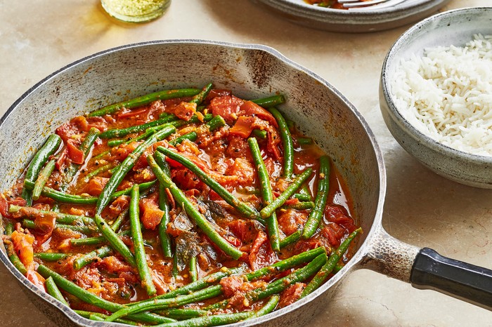 a metal pan containing green beans and tomato with a bowl of rice on a stone background