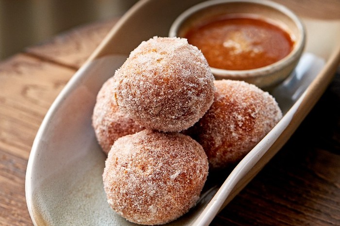 A white oval dish filled with sugar-coated doughnuts and a pot of sauce on a wooden table