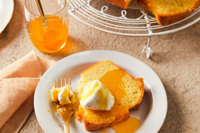 An orange drizzle cake loaf on a cake stand with a slice of cake on a separate plate topped with a dollop of cream