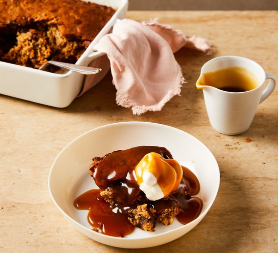 A bowl of sticky pudding topped with a dollop of cream, plus a jug of toffee sauce and a baking tray of sticky toffee pudding