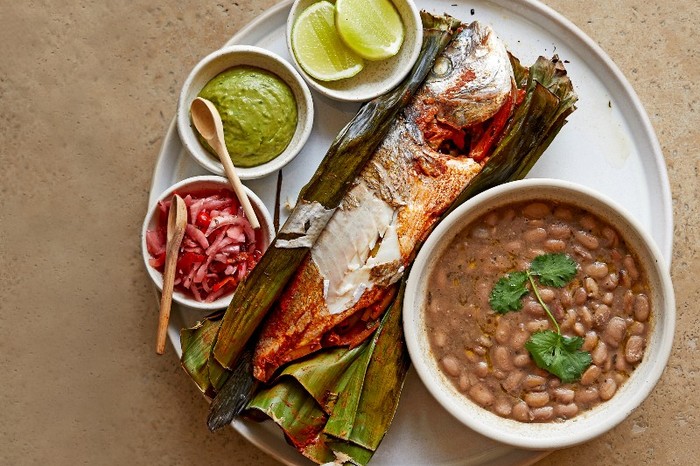 A plate filled with a whole seabream cooked in a banana leaf and various small bowls of sauces and chutneys on a wooden background